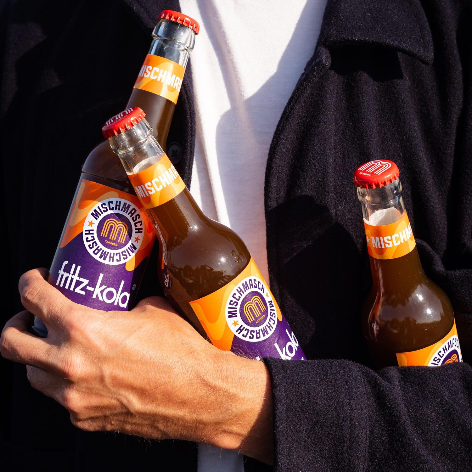 Eine Person in schwarzem Hemd hält drei Flaschen fritz-kola Mischmasch in den Armen. Die Flaschen zeigen das charakteristische orangefarbene und violette Mischmasch-Design mit dem 'M' Logo. Das Bild ist in starkem Sonnenlicht aufgenommen, das einen deutlichen Kontrast zwischen hell und dunkel erzeugt.