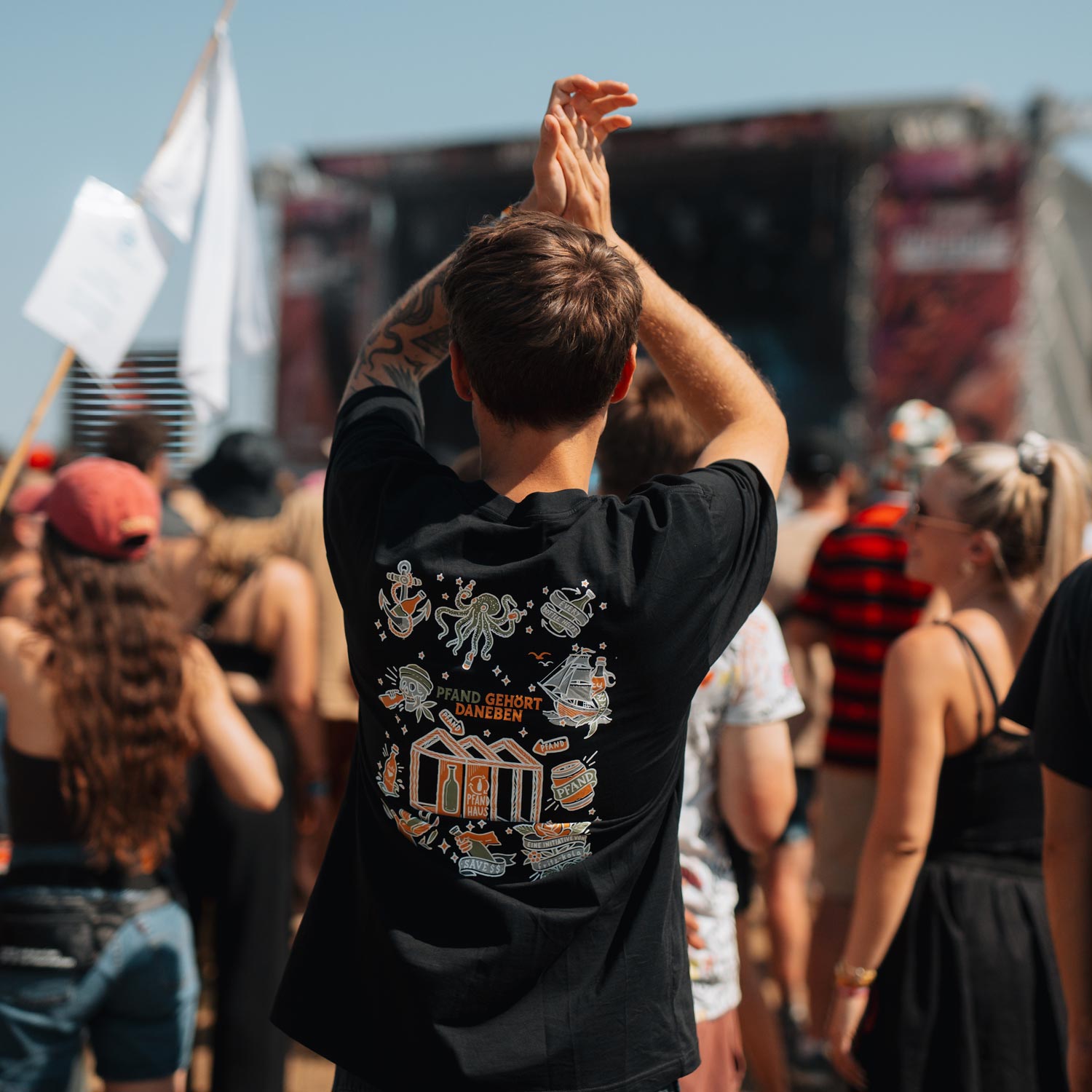 Ein Mann auf einem Festival reckt jubelnd die Arme in die Höhe, während er das schwarze T-Shirt der "Pfand gehört daneben"-Kollektion trägt. Das Rückendesign mit maritimen Motiven und dem Pfandhaus ist deutlich sichtbar. Im Hintergrund sind Festivalgäste und eine Bühne zu erkennen.