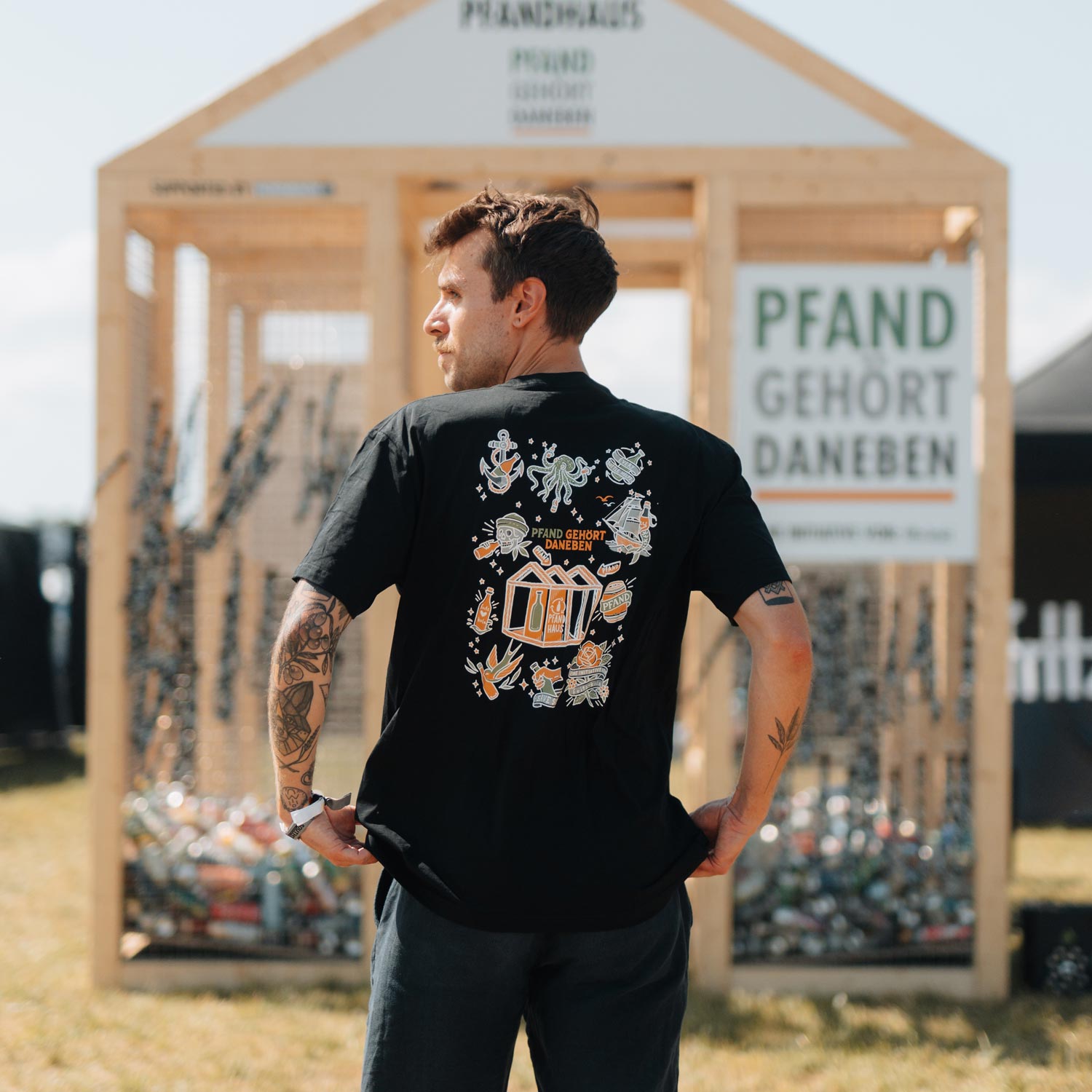 Ein tätowierter Mann im schwarzen T-Shirt der Kollektion steht vor dem Pfandhaus und blickt seitlich. Sein Rücken zeigt das kunstvolle Design mit maritimen Elementen und dem zentralen Pfandhaus-Motiv. Im Hintergrund sind weitere Festivalstände und das fritz-kola Logo erkennbar.