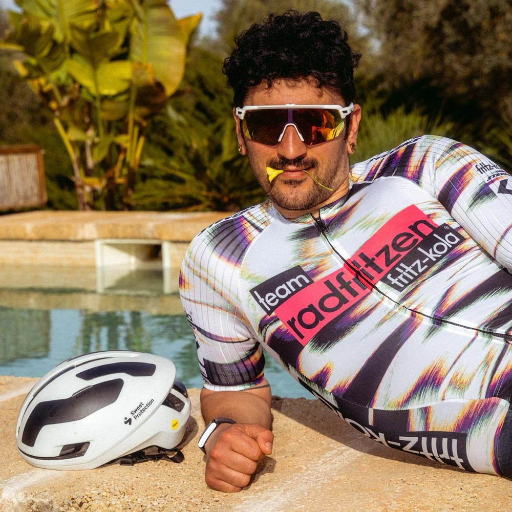 Radsportler mit lockigen Haaren und bunter Sonnenbrille posiert am Poolrand. Er trägt das Team radfritzen fritz-kola Trikot mit charakteristischen bunten abstrakten Streifen. Weißer Helm liegt neben ihm, türkisfarbenes Wasser im Hintergrund.