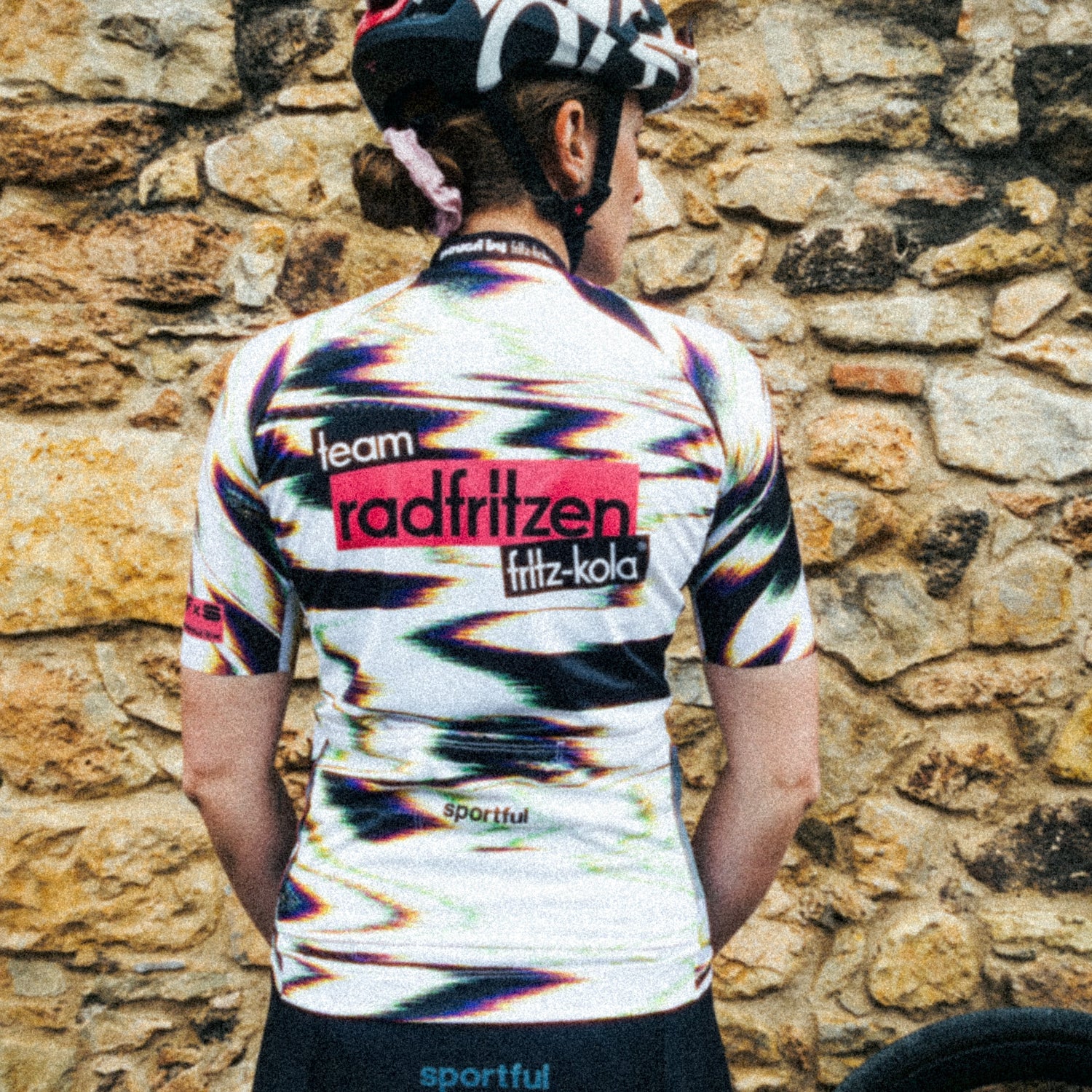 Rückenansicht des radfritzen Trikots mit großem „team radfritzen fritz-kola“ Schriftzug und dynamischem Farbdesign, Fahrrad im Hintergrund.
