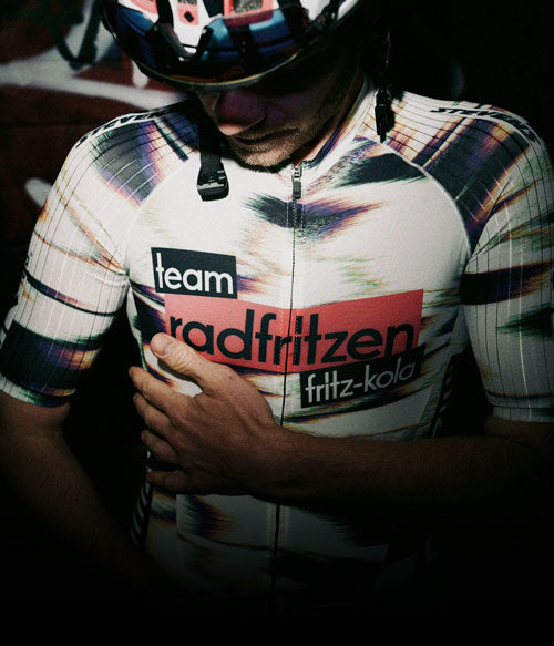 Nahaufnahme einer Person in Fahrradkleidung und Helm. Auf dem Trikot steht „team radfritzen“ sowie „fritz-kola“. Eine Hand liegt auf der Brust, der Hintergrund ist dunkel und unscharf.