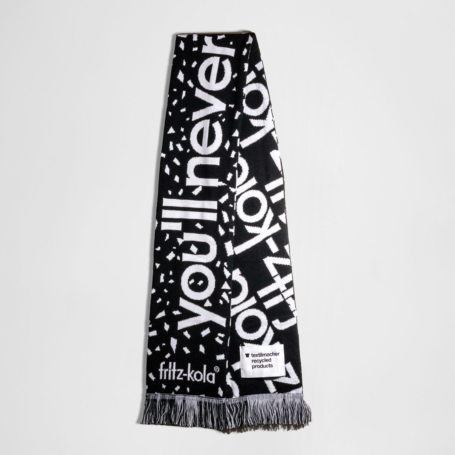 ein schwarz-weißer Fanschal im Design von fritz-kola. Der Schal zeigt den Text 'You'll never drink alone'  auf schwarzem Hintergrund mit weißer Schrift auf der einen Seite. Auf der anderen Seite wiederholt sich auf schwarzem Hintergrund in weißer Schrift der Text "fritz-kola". Am unteren Ende befinden sich schwarz-weiße Fransen.
