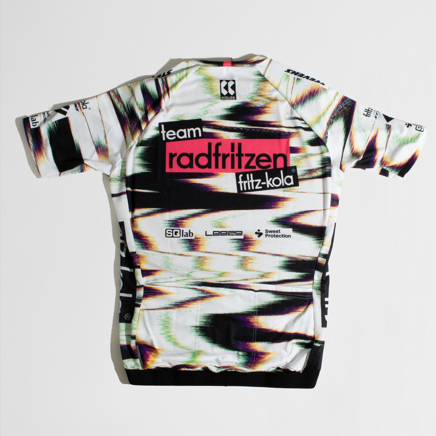 Rückseite Weißes Radtrikot mit abstraktem Farbverlauf-Muster in Grün, Pink und Schwarz. Zentraler roter Balken mit "radfritzen" auf der Brust, darunter  "fritz-kola" Logo. Schwarze Seitenstreifen mit fritz-kola Schriftzug, kurze Ärmel, Reißverschluß