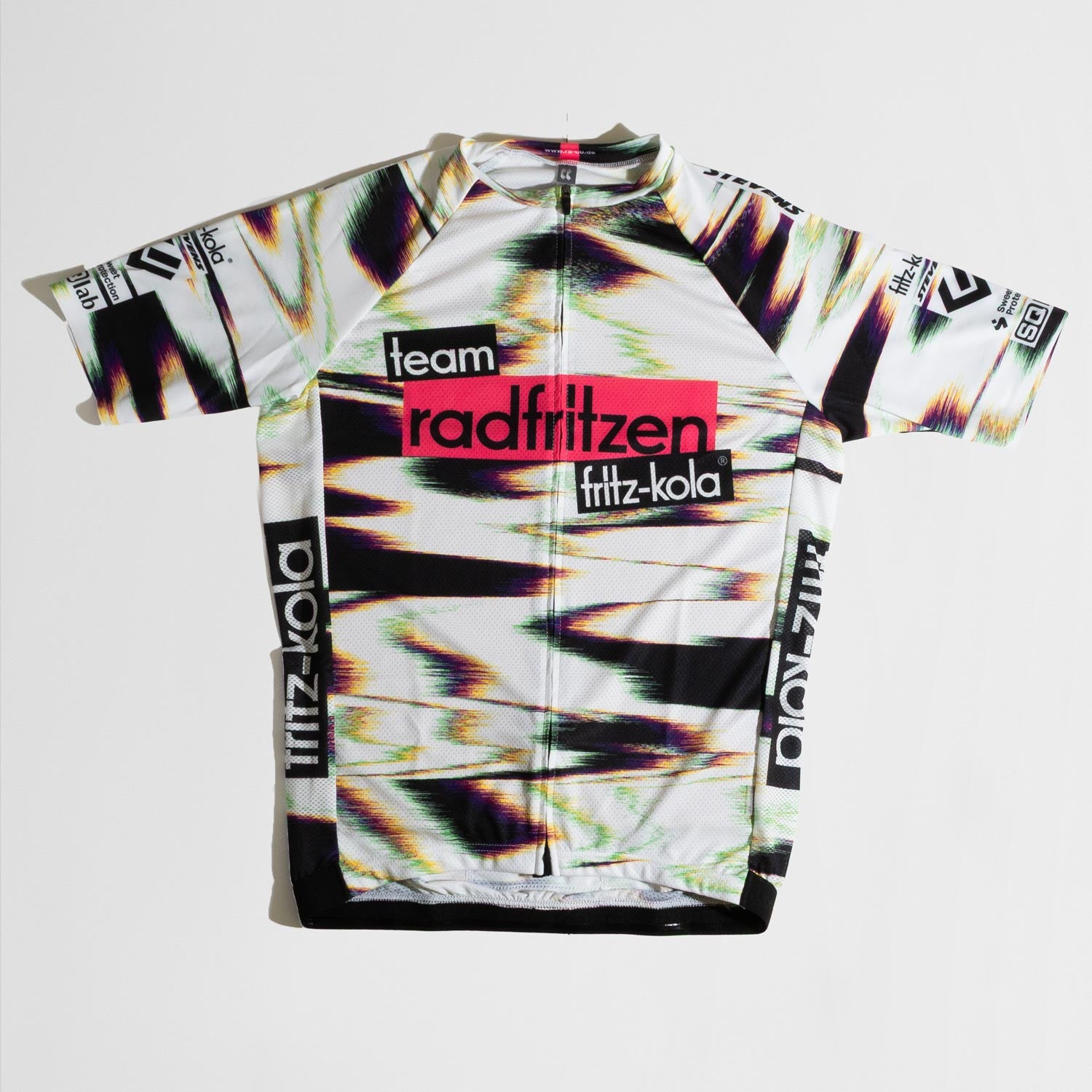 Weißes Radtrikot mit abstraktem Farbverlauf-Muster in Grün, Pink und Schwarz. Zentraler roter Balken mit "radfritzen" auf der Brust, darunter  "fritz-kola" Logo. Schwarze Seitenstreifen mit fritz-kola Schriftzug, kurze Ärmel, Reißverschluß