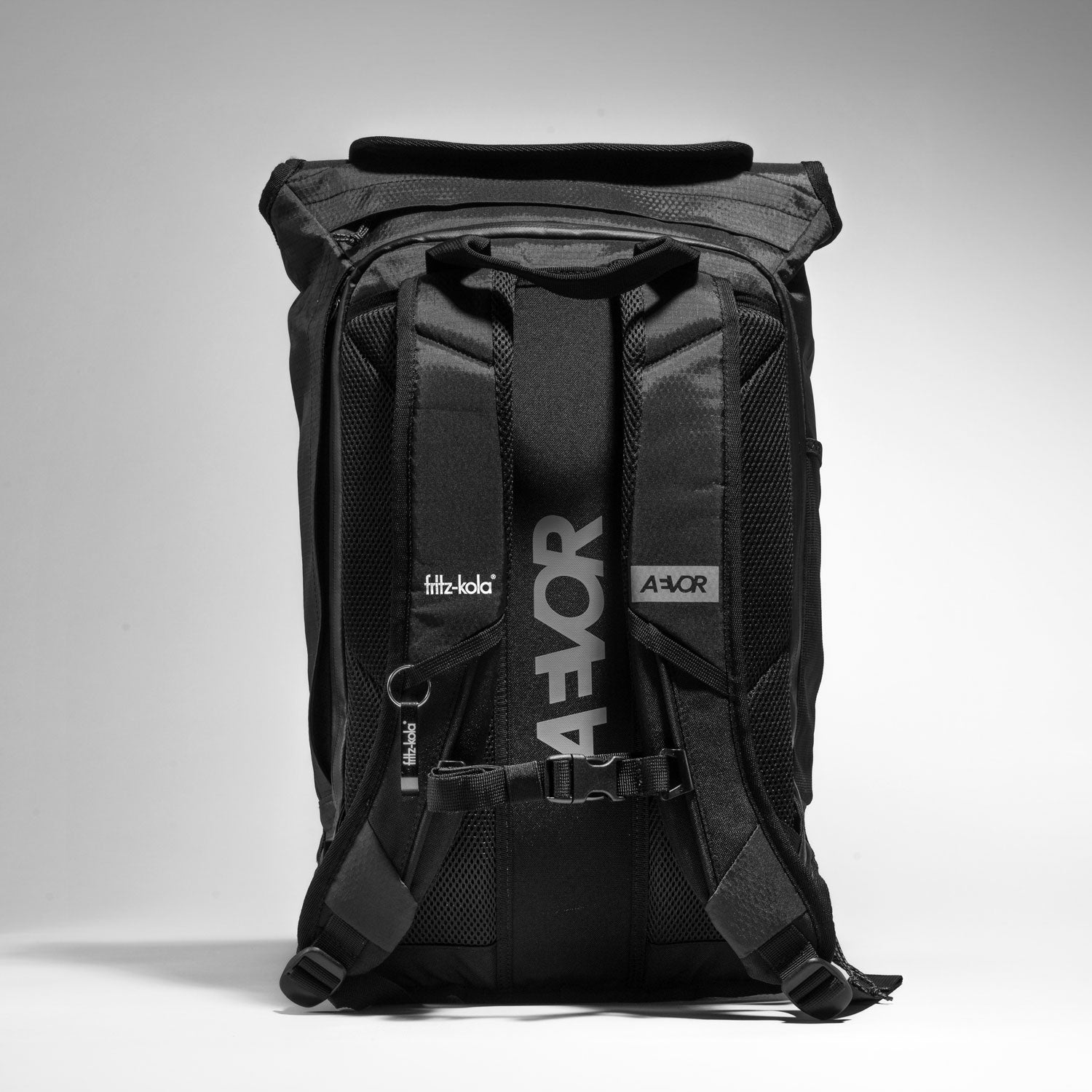Der schwarze Aevor trip pack Rucksack in der limitierten fritz-kola edition von hinten vor hellem hintergrund. Es ist an dem rechten Schultergurt ein Flaschenöffner angehangen und das fritz-kola Logo ist drauf gedruckt.
