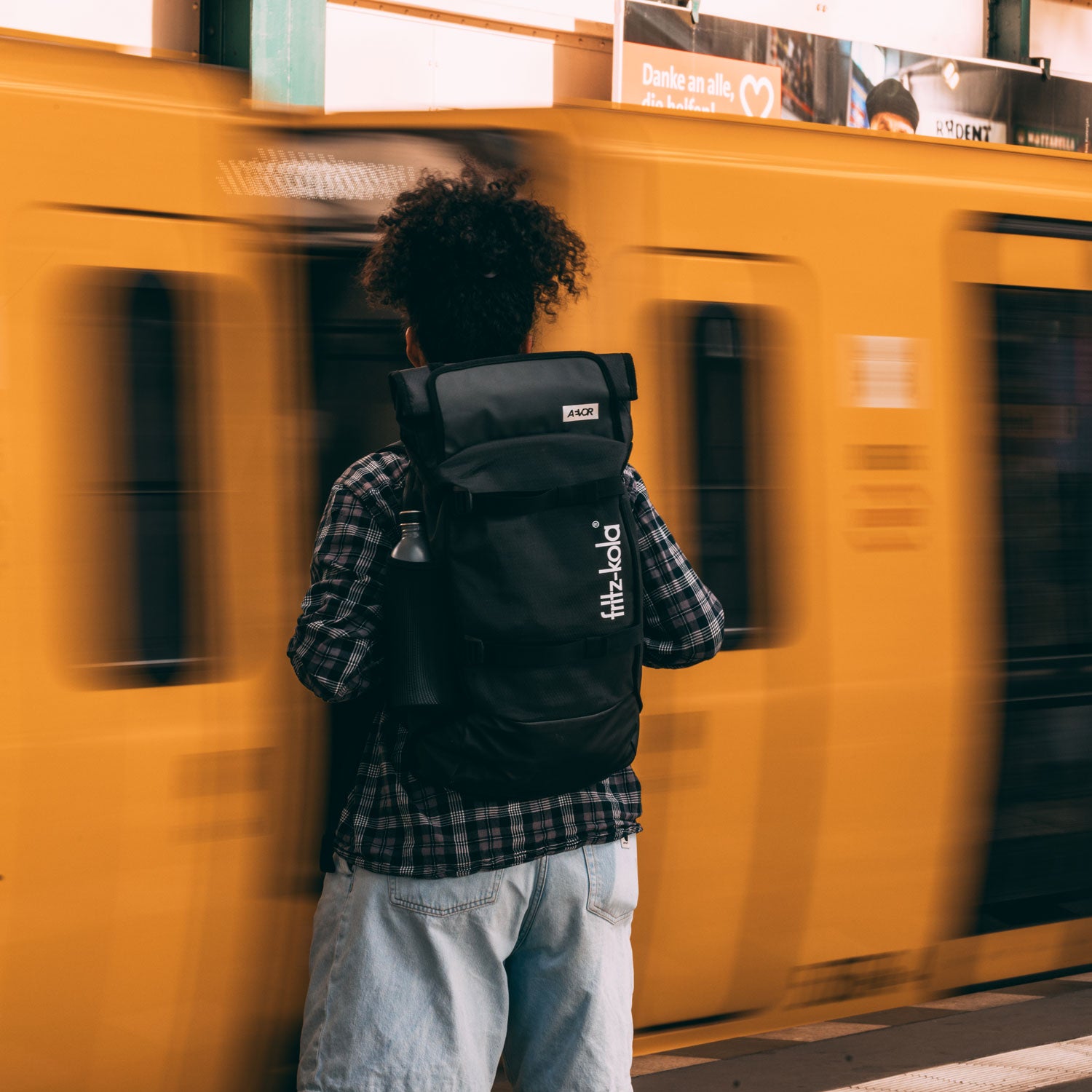 Eine Person mit einem schwarzen fritz-kola Rucksack steht vor einer vorbeifahrenden gelben U-Bahn. Sie trägt ein kariertes Hemd und helle Jeans-Shorts. Der Bewegungseffekt der U-Bahn ist durch Bewegungsunschärfe dargestellt.