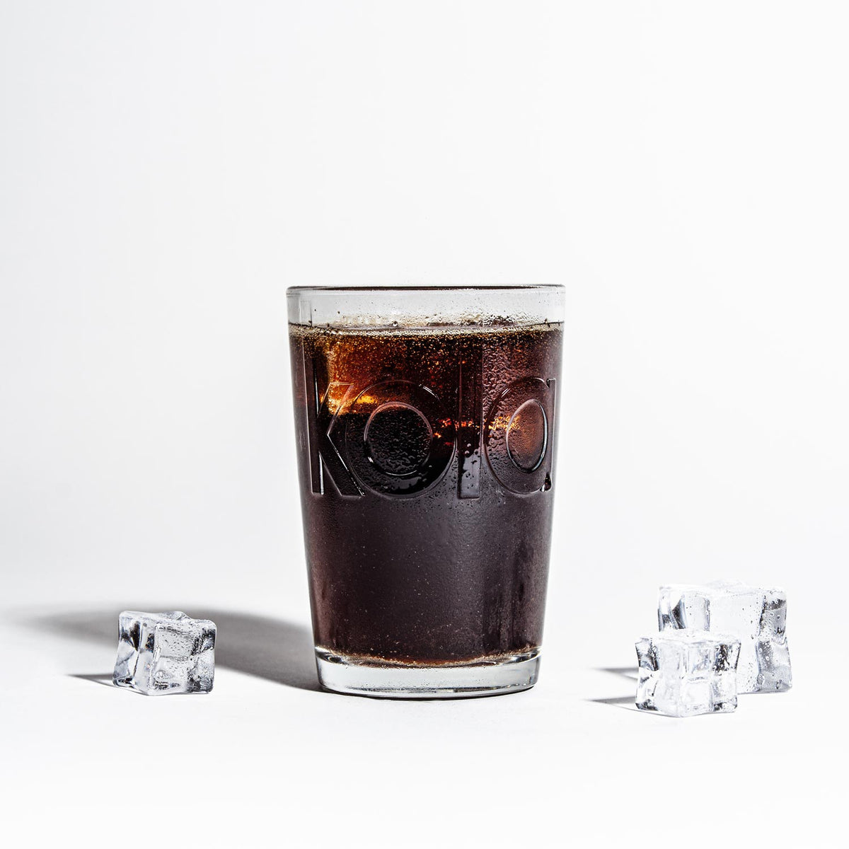 Ein mit fritz-kola gefülltes Glas, flankiert von Eiswürfeln auf beiden Seiten. Die dunkle Cola kontrastiert stark mit dem weißen Hintergrund. Der eingeprägte kola  Schriftzug ist deutlich sichtbar, die Oberfläche des Glases zeigt Kondensation. 