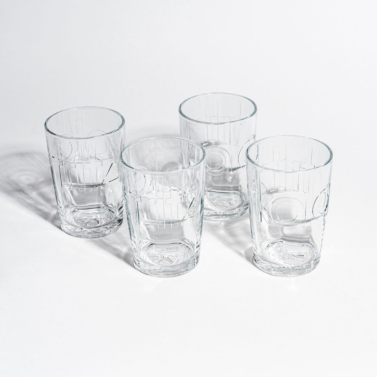 Vier leere fritz-kola Gläser auf weißem Hintergrund, fotografiert von oben. Die Gläser sind in quadratischer Anordnung platziert und zeigen deutlich die eingeprägten fritz-kola Schriftzüge. Klares Kristallglas mit charakteristischen Rillen und Prägungen wirft dezente Schatten.