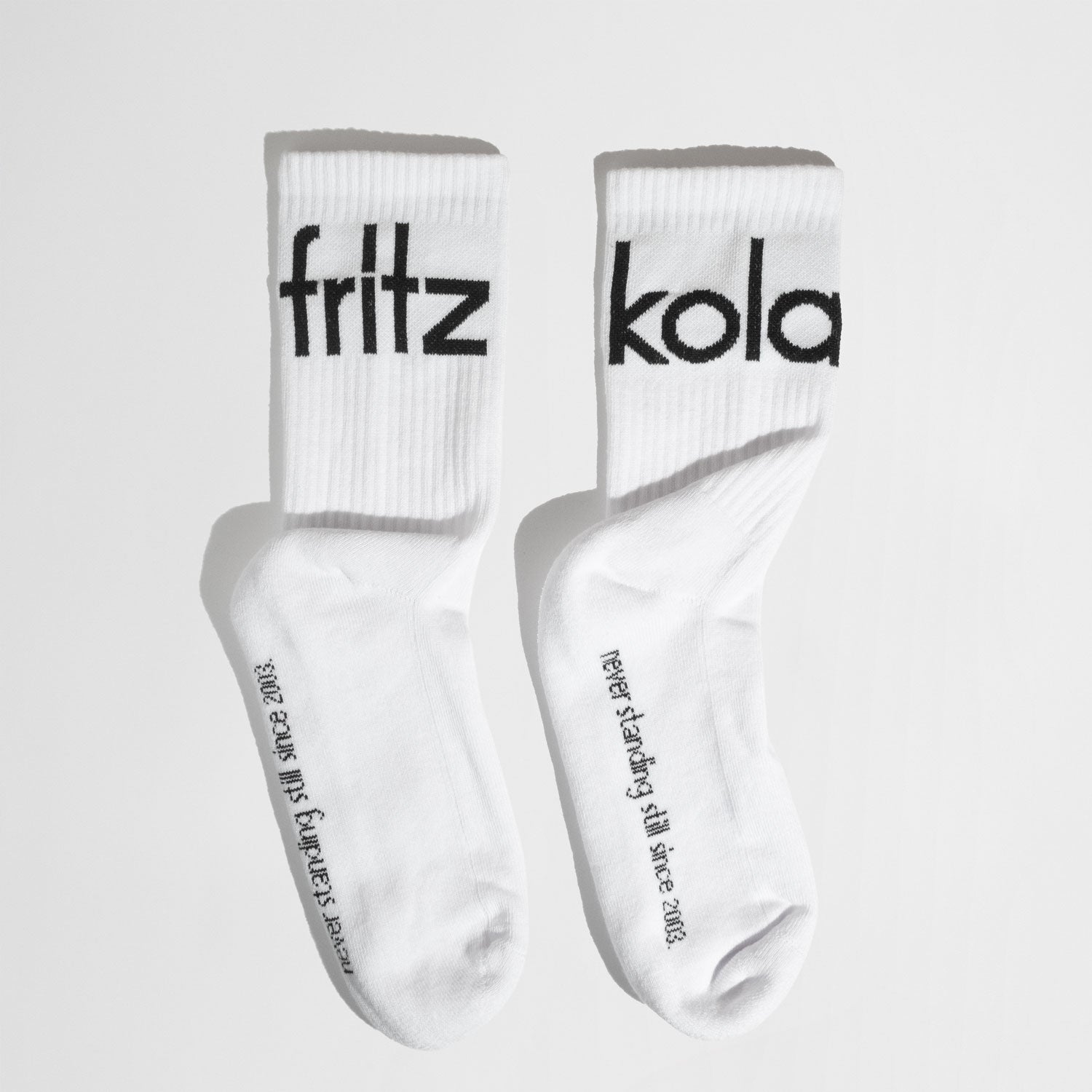 Ein Paar weiße Socken mit schwarzem Stick. Auf der linken Socke steht „fritz“, auf der rechten „kola“. Auf dem Fußteil befindet sich zusätzlich der Text „Never standing still since 2003"