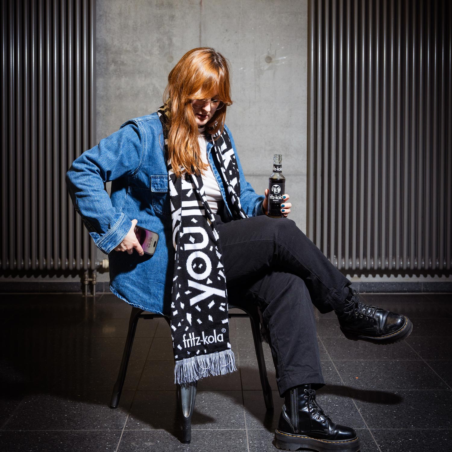 Das Bild zeigt eine Person in lässiger Streetwear mit einem fritz-kola Schal. Sie trägt eine Jeansjacke, schwarze Hose und Stiefel. In der Hand hält sie eine Flasche fritz-kola. Der schwarz-weiße Schal mit dem fritz-kola Logo und geometrischen Mustern hat Fransen an den Enden