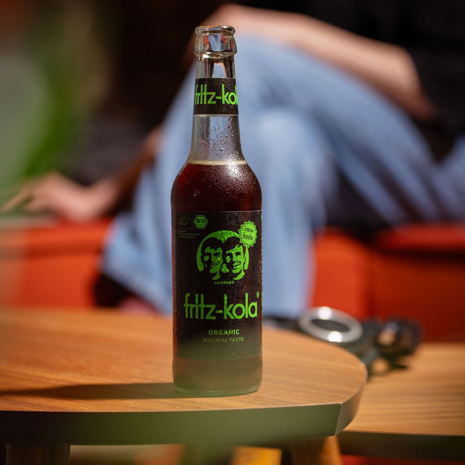 Glasflasche fritz-kola Organic Bio-Kola steht auf einem Tisch, im Hintergrund eine unscharfe sitzende Person