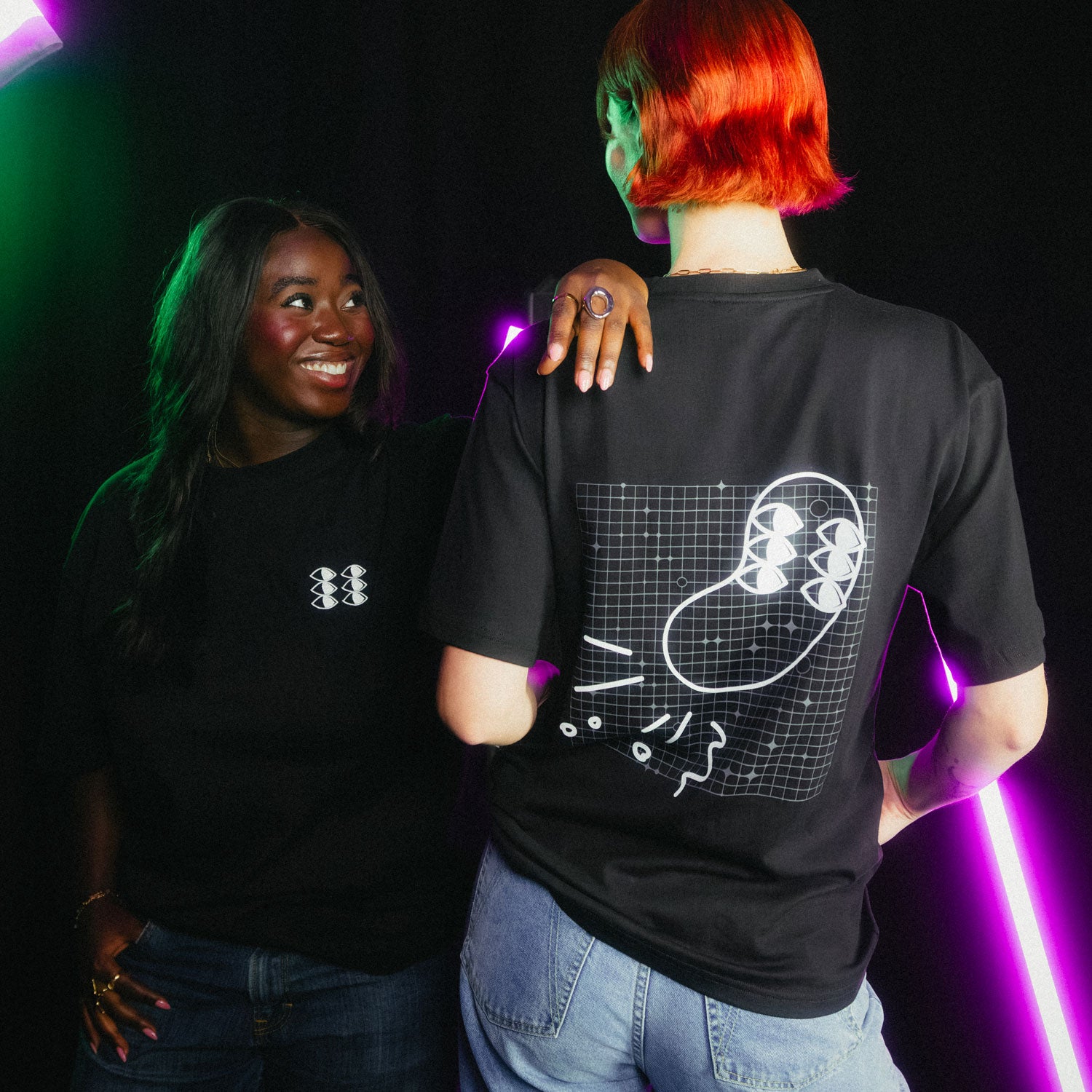 Zwei Personen in schwarzen fritz-kola x rktbns T-Shirts unter violett-grünem Neonlicht - links lächelnde Frau mit kleinem Brustlogo, rechts Person mit kupferfarbenen Haaren und großem Rückenprint