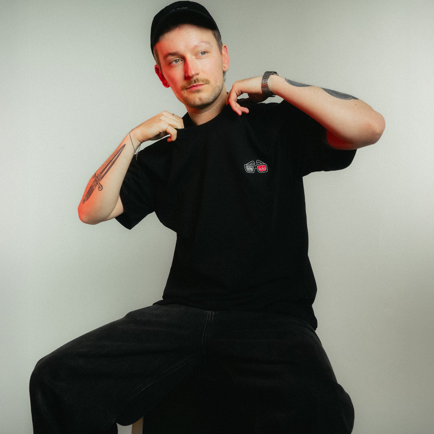 Mann mit schwarzer Cap und Armtattoos in schwarzem fritz-kola x rktbns T-Shirt mit kleinem Brustlogo, beide Hände am Kragen in lässiger Pose vor hellem Hintergrund. Brustlogo stellt eine 3d Brille mit fritz-kola Schriftzug dar