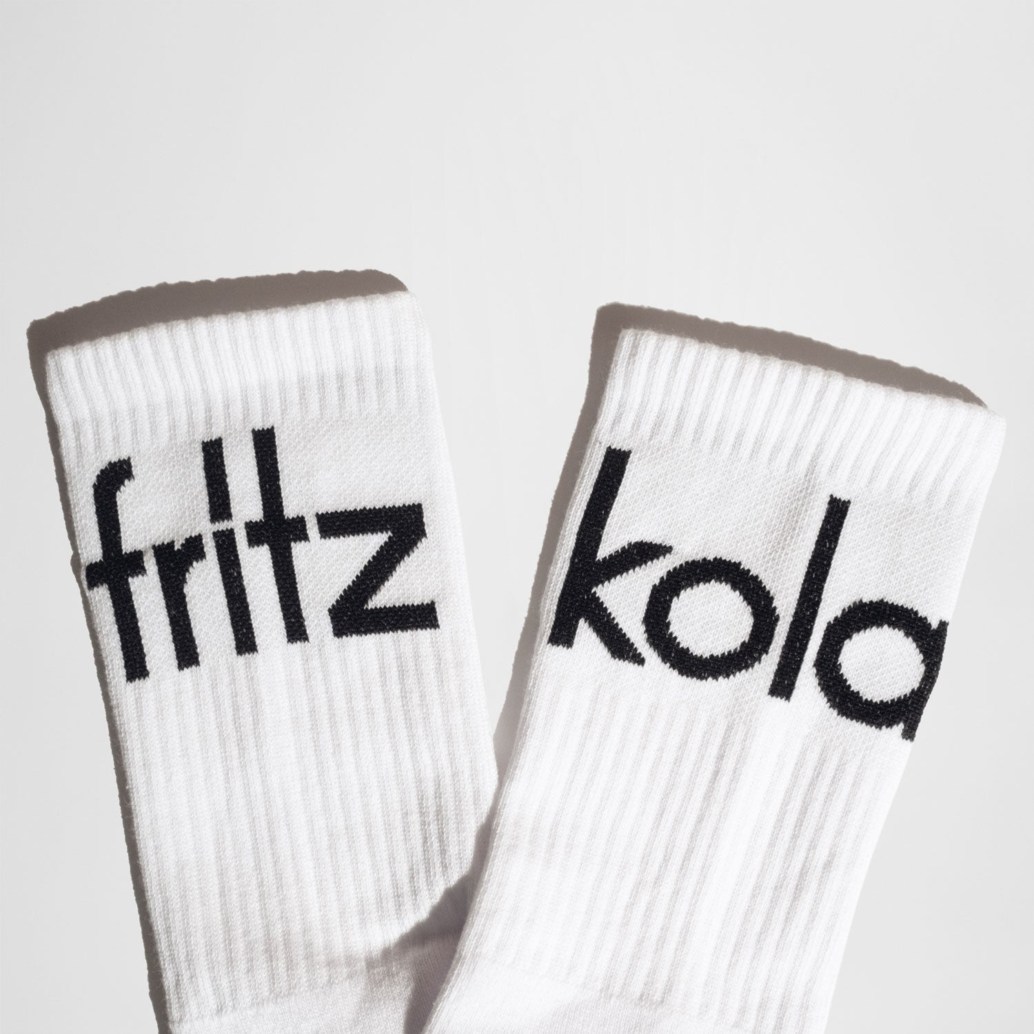 Ein Paar weiße Socken mit schwarzem Stick. Auf der linken Socke steht „fritz“, auf der rechten „kola“. Die Socken sind Teil des fritz-kola Merchandise und zeigen das markante Branding.