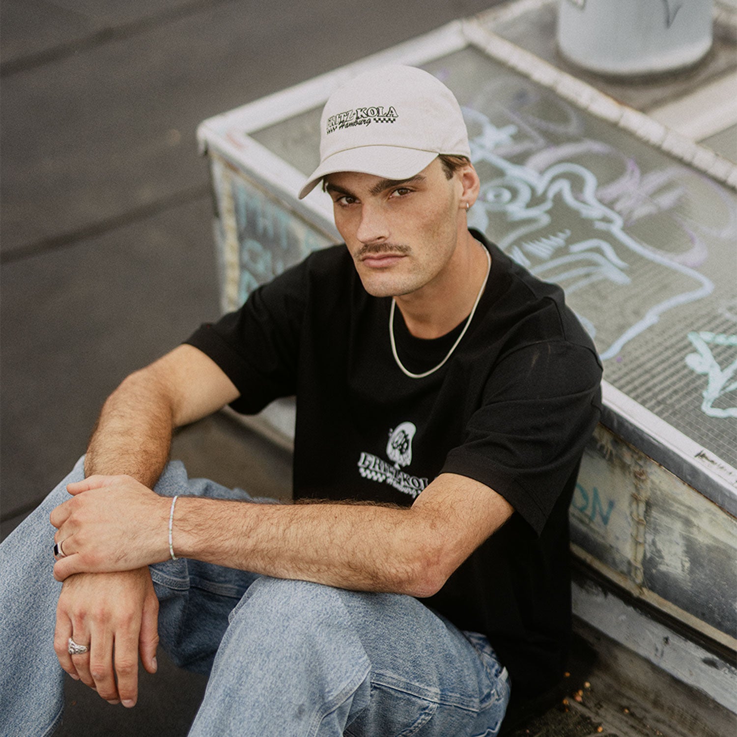 Mann mit Schnurrbart trägt beige fritz-kola Cap und schwarzes T-Shirt mit fritz-kola Logo. Sitzt entspannt in blauen Jeans vor Graffiti-Wand, goldene Kette um den Hals, Silberarmband am Handgelenk.