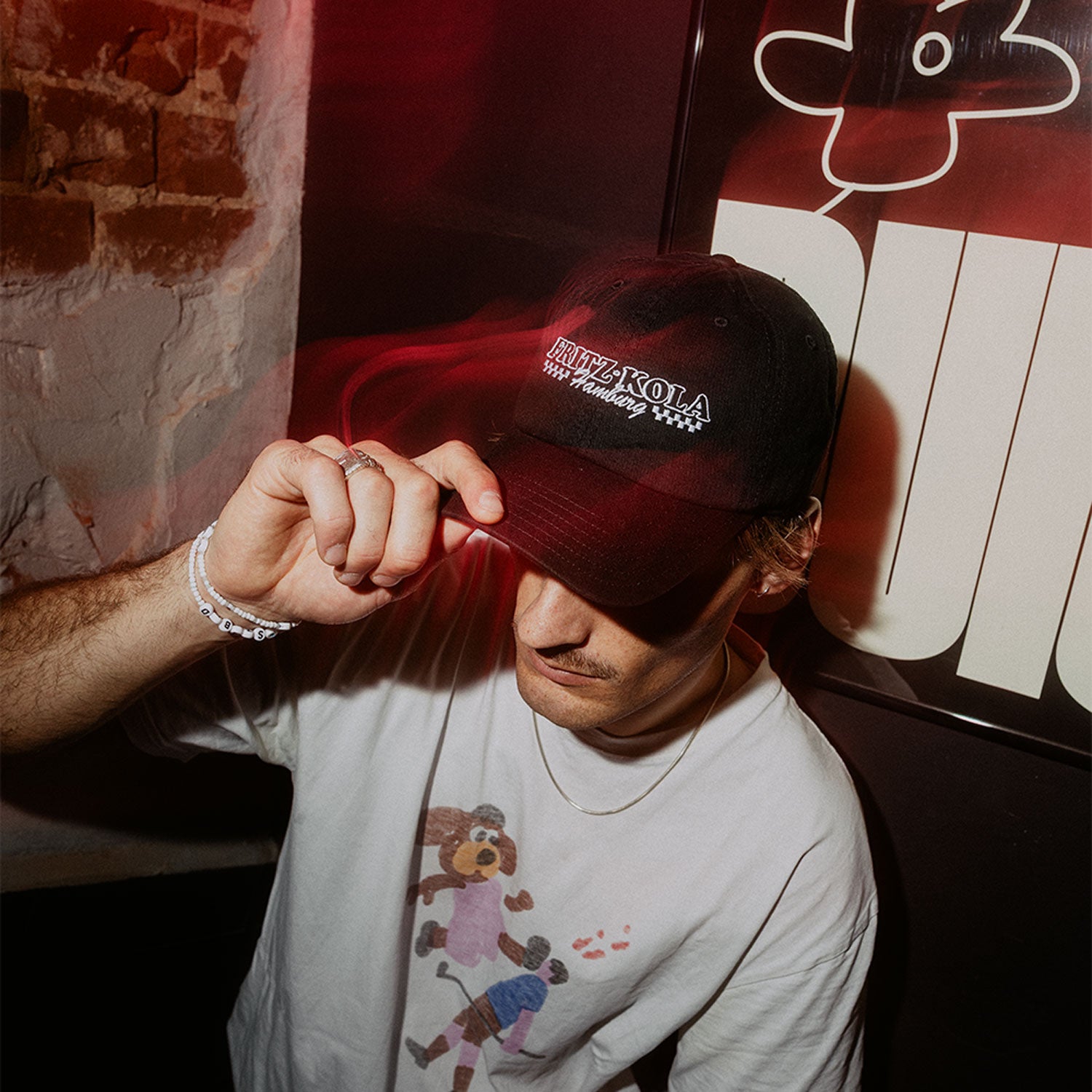 Mann in weißem Shirt  trägt schwarze fritz-kola Cap. Fasst mit der Hand an den Schirm, warmes rotes Licht beleuchtet die Szene vor Backsteinwand.