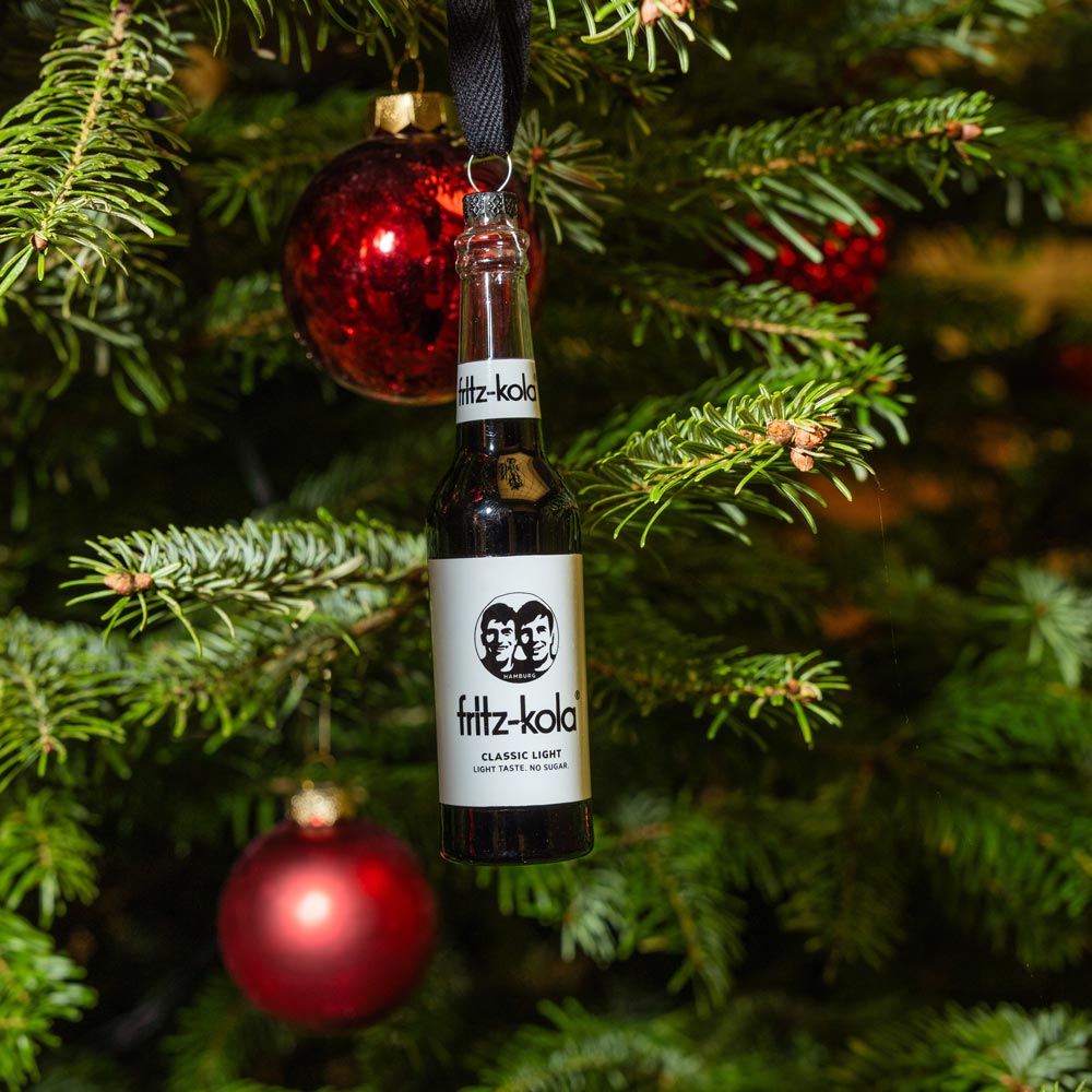 Eine fritz-kola classic light Flasche hängt an einem schwarzen Band am Weihnachtsbaum. Im Hintergrund sind rote Christbaumkugeln und grüne Tannenzweige zu sehen.