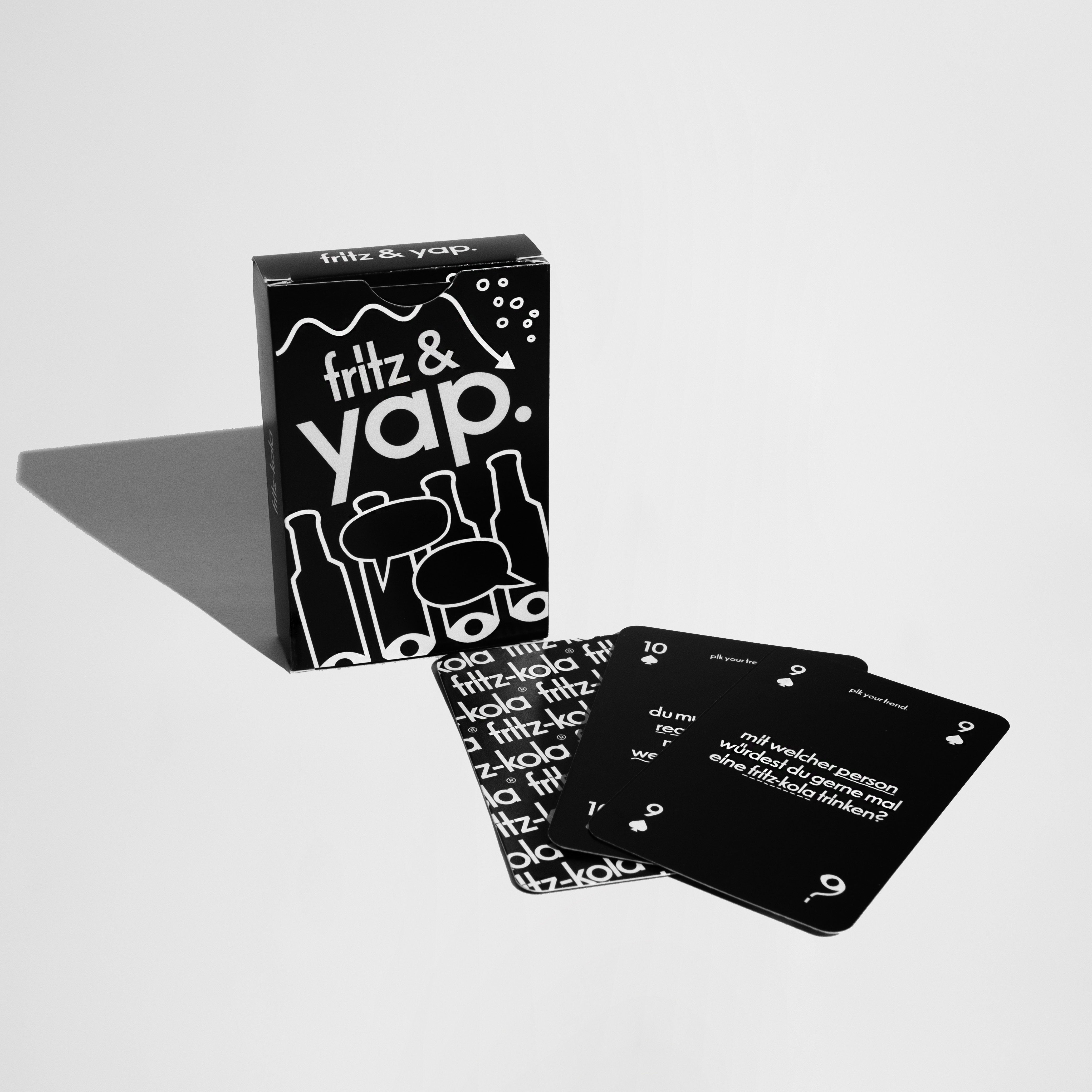 Schwarze Spielkarten und eine Spielbox mit der Aufschrift ‚fritz & yap.‘ in weißer Schrift. Die Box zeigt stilisierte Flaschen und Gläser, während die Karten mit weißen Texten und Zahlen bedruckt sind. Das Set liegt auf einer hellen Fläche mit Schattenwurf.