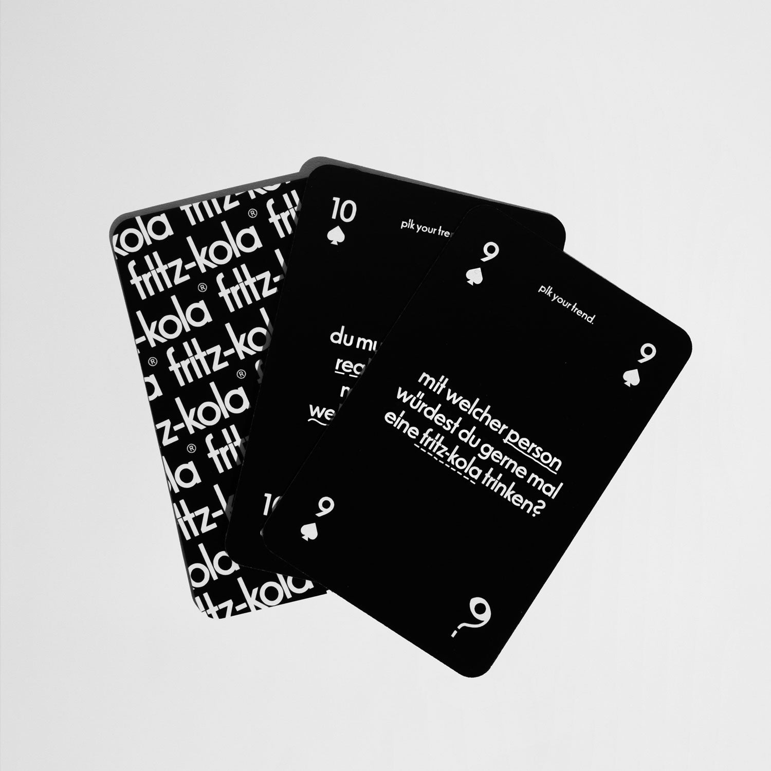 Vier schwarze Spielkarten aus dem ‚fritz & yap‘-Set, teilweise überlappend. Die Vorderseiten zeigen weiße Schrift mit Fragen und Zahlen, darunter eine Karte mit der Frage: ‚Mit welcher Person würdest du gerne mal eine fritz-kola trinken?‘. Die Rückseite einer Karte ist mit dem fritz-kola-Logo-Muster bedruckt.