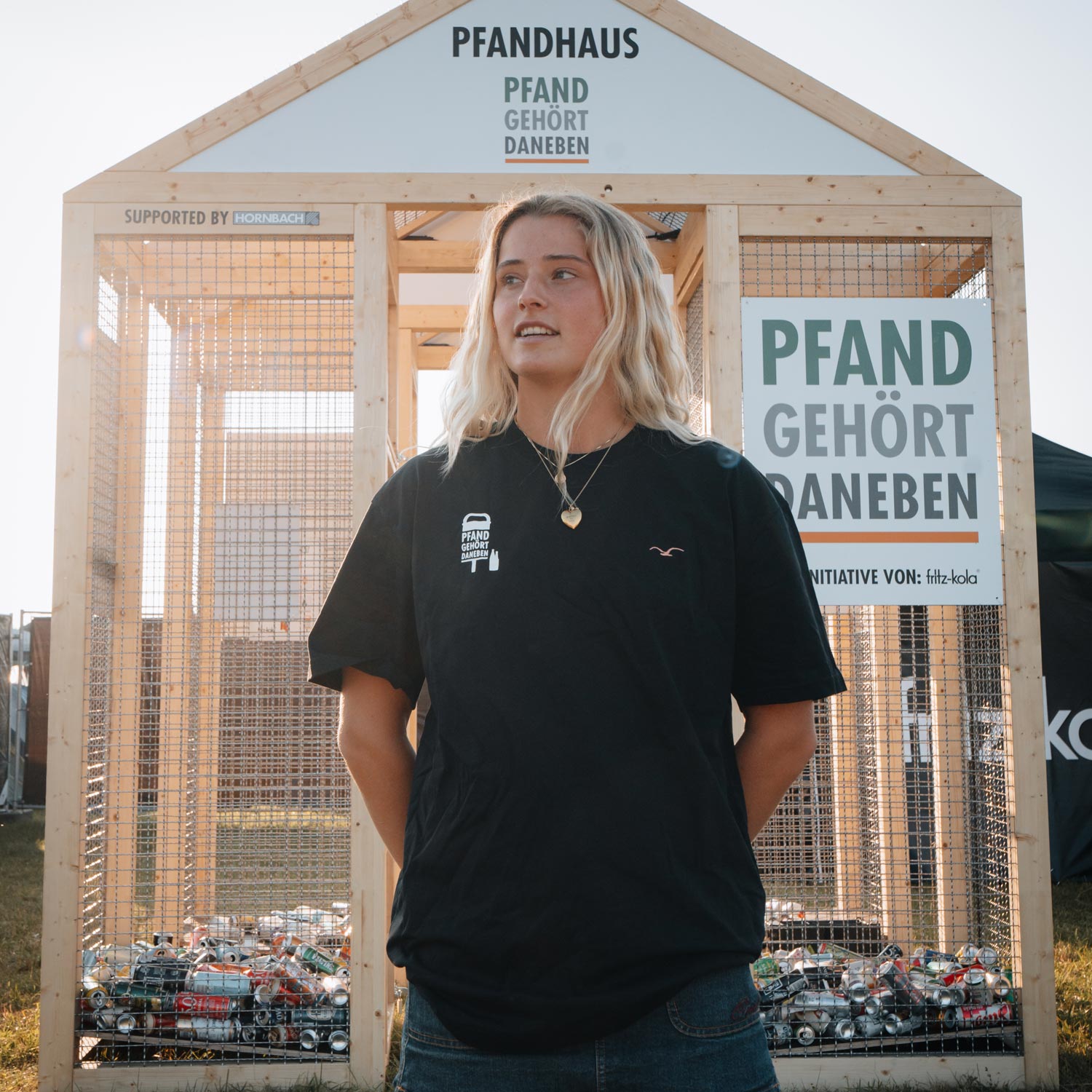 Eine blonde Frau im schwarzen T-Shirt der Kollektion steht entspannt vor dem Pfandhaus. Sie trägt einen Jeansrock und eine Halskette. Das Pfandhaus im Hintergrund ist mit bunten Pfandflaschen gefüllt und zeigt das "PFAND GEHÖRT DANEBEN"-Schild der fritz-kola Initiative.