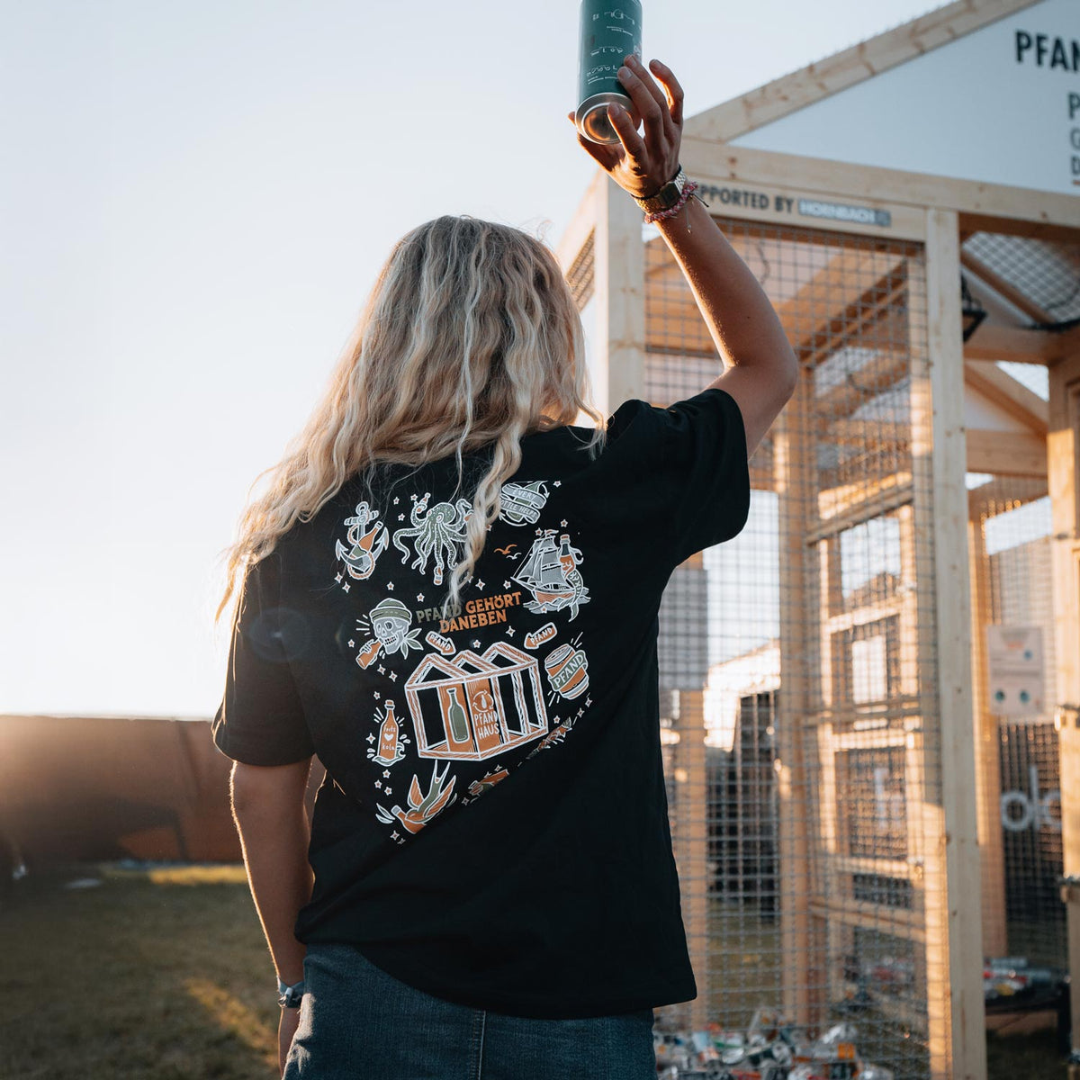 Eine Person mit blonden Haaren wirft eine Pfanddose ins Pfandhaus, während sie das schwarze Shirt der "Pfand gehört daneben"-Kollektion trägt. Das detailreiche Rückendesign mit Pfandhaus-Motiv ist im warmen Abendlicht zu sehen. Im Hintergrund steht das hölzerne Pfandhaus mit gesammelten Flaschen.