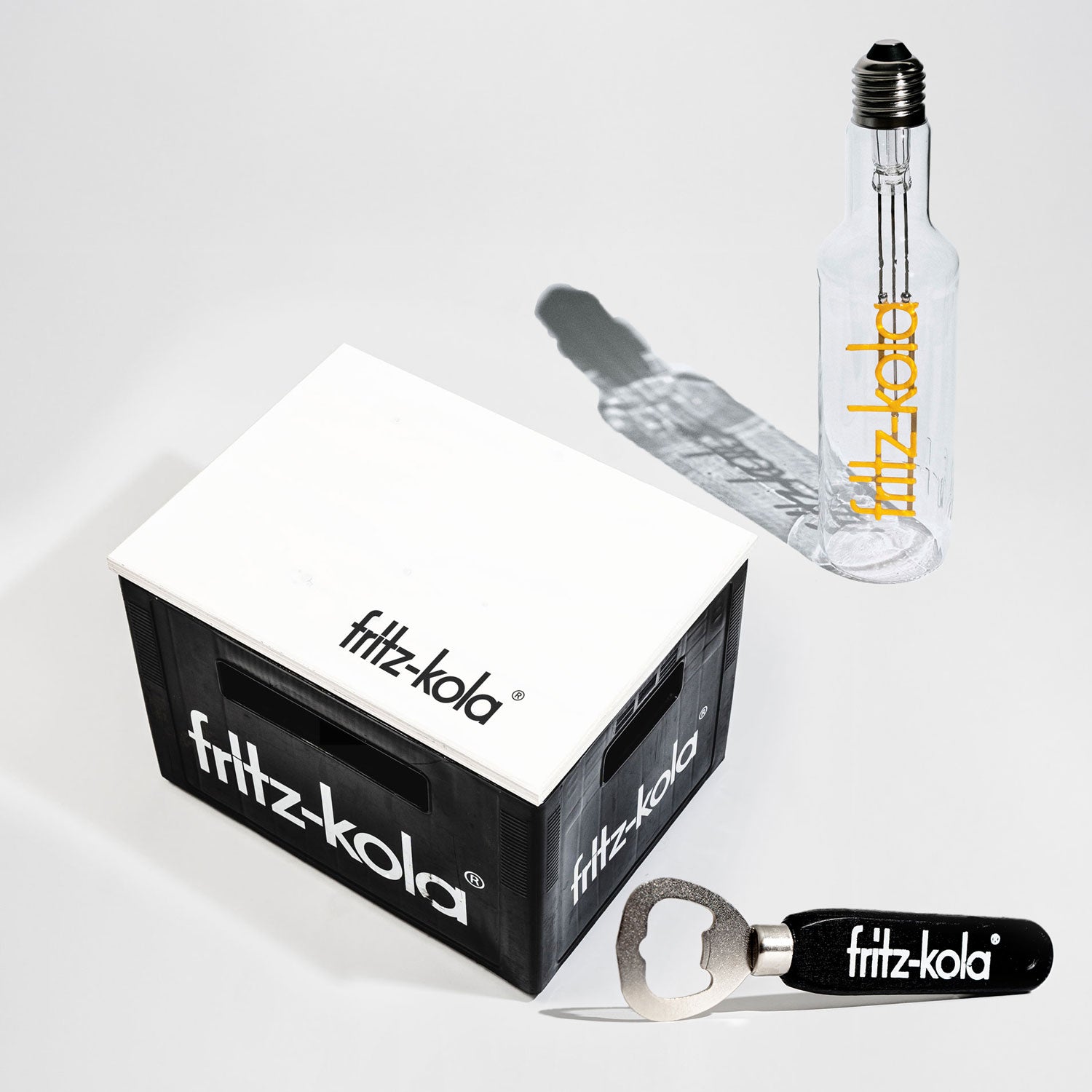 Set aus fritz-kola Merchandise für Zuhause: schwarze Mini-Kiste mit weißem fritz-kola-Logo, transparente Glasflasche im Glühbirnen-Design mit gelbem fritz-kola-Schriftzug und ein schwarzer Flaschenöffner mit weißem fritz-kola-Logo.